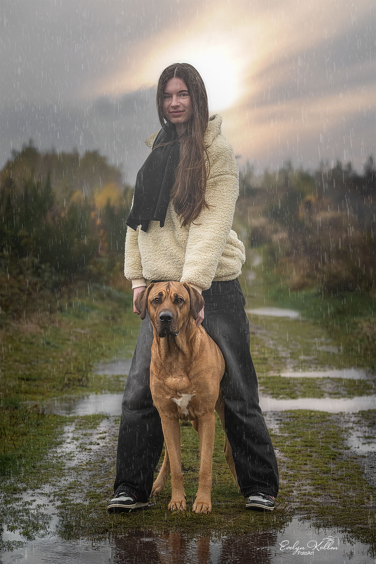 Woman in a cream fleece jacket and black scarf stands in a rainy field holding a large brown dog, sun halo behind them.