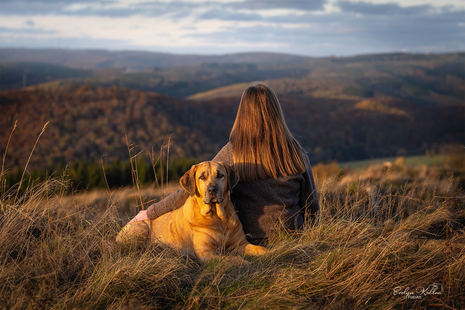 Woman with long hair sits in tall grass on a hill at sunset, hugging a dog as a valley stretches into the distance.