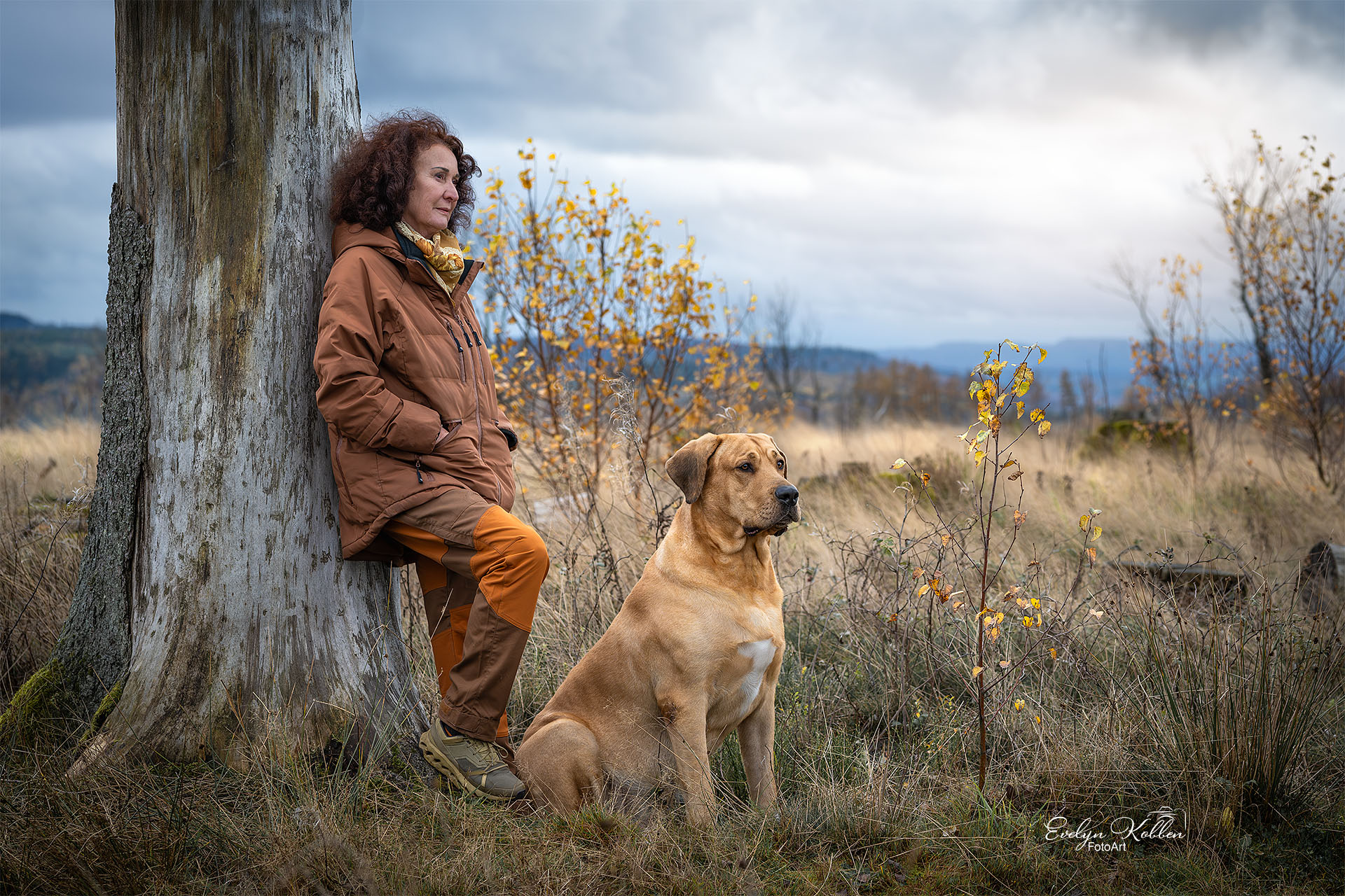 Hundefotografie - Eine Frau genießt mit ihrem Broholmer die Natur, lehnt an einem Baum